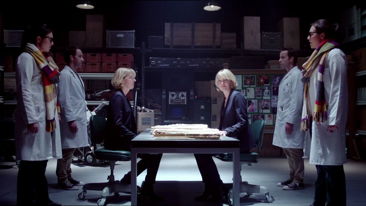 An image from the Doctor Who episode "The Day of the Doctor", showing human and Zygon Kate Stewarts, and their assistants, facing off on either side of a table.